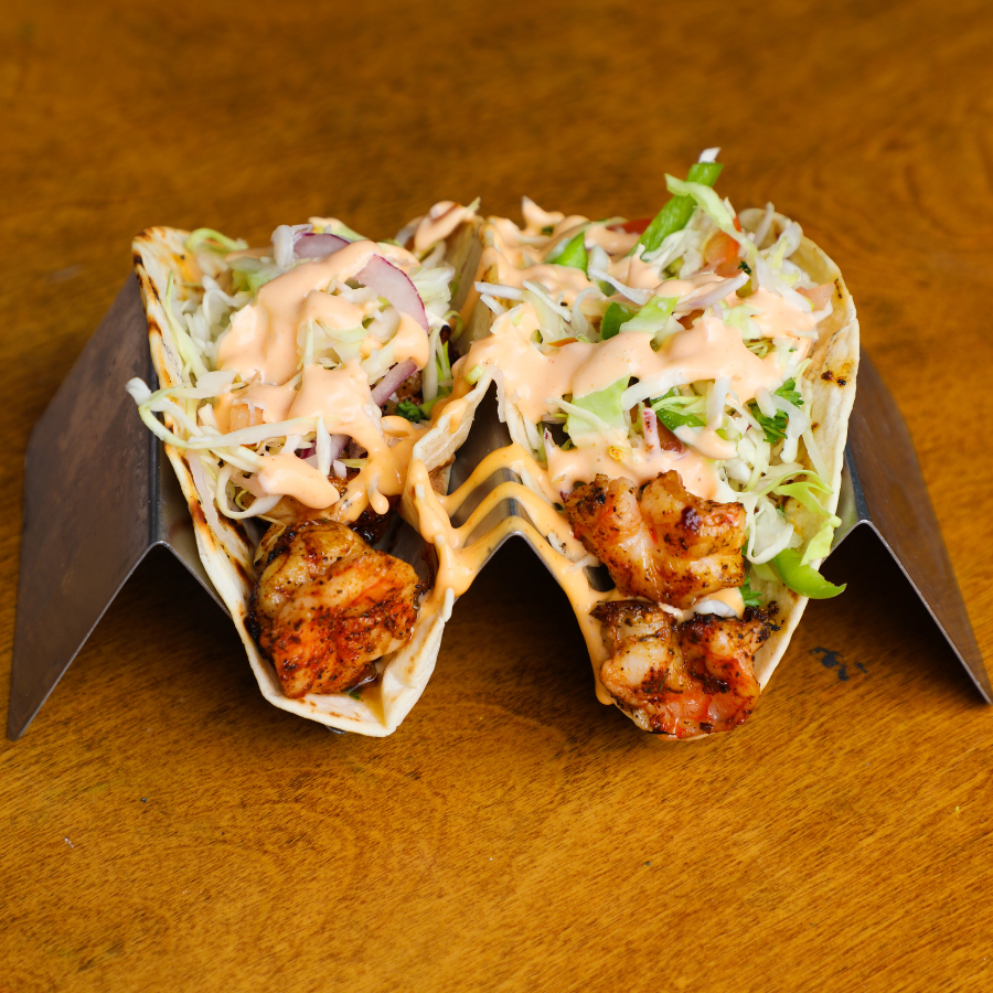 Two shrimp tacos on a silver taco holder on a wooden table.